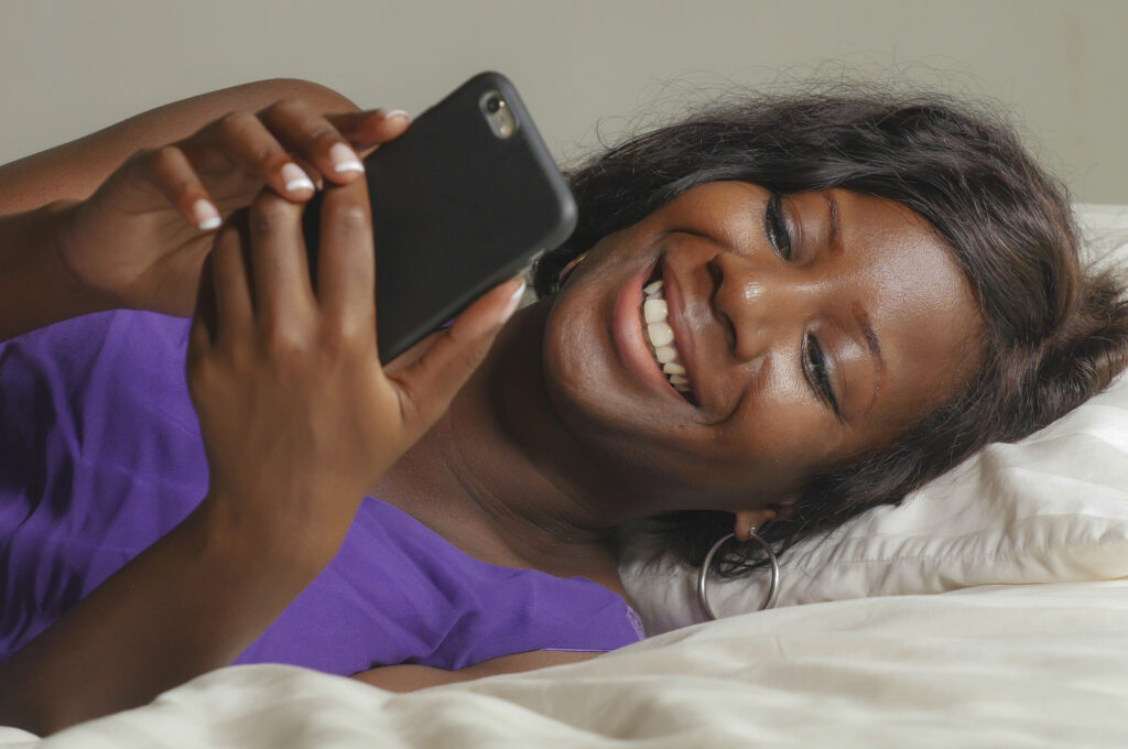 African woman lying on bed smiling at smart phone.